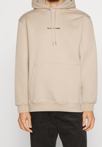 Felpa beige con cappuccio, coulisse, tasca frontale e piccolo logo nero sul petto. Presenta una vestibilità rilassata e un materiale morbido in misto cotone.