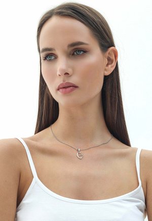 Collana d'argento con un pendente a forma di cuore su una catena sottile, esposta su uno sfondo semplice. La modella indossa un top bianco senza maniche.