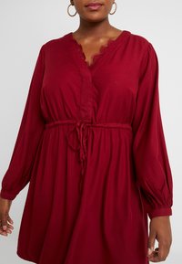 Abito rosso con maniche lunghe e scollatura a V. Presenta un bordo in pizzo e un nastro in vita per la regolazione. Tessuto morbido con vestibilità ampia e maniche a palloncino.
