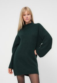 Tumši zaļš pārāk liels adīts džemperis ar augstu apkakli, garām piedurknēm un teksturētu rakstu, kas krīt virs ceļa.