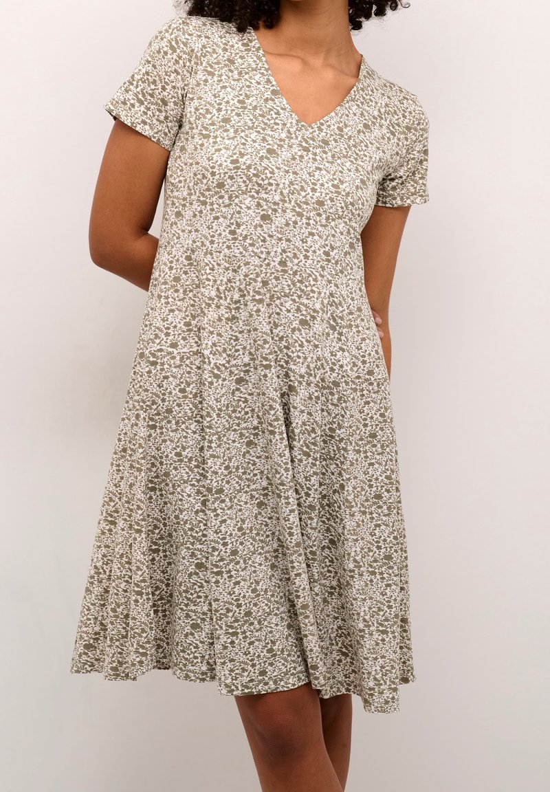 Femme portant une robe à manches courtes, col en V, longueur genoux avec un motif floral beige et blanc, debout devant un fond clair uni.