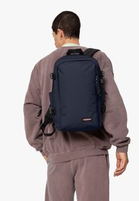 Zaino navy con forma rettangolare, realizzato in tessuto resistente, presenta spallacci regolabili, fibbia frontale e una piccola etichetta con logo.