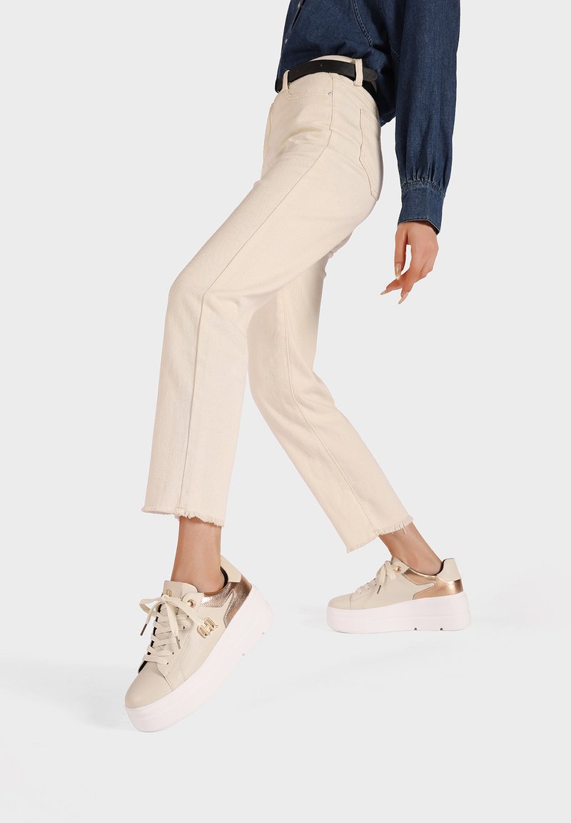 Jeans de talle alto en color beige con dobladillo deshilachado, combinados con zapatillas crema que tienen acentos metálicos dorados y una suela gruesa blanca.