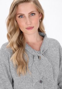 Cardigan gri din tricot cu închidere cu cravată la guler, accente de butoni argintii și o textură moale. Prezintă o croială lejeră și un aspect confortabil.
