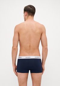Morski plavi bokserice s bijelim elastičnim pojasom na kojem se nalazi logo "Lacoste". Materijal izgleda glatko i prilagođeno, dizajniran za udobnost.