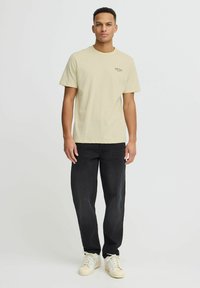 Camiseta de algodón beige con mangas cortas y cuello redondo. Presenta un pequeño logo de texto negro en el lado izquierdo. Combinada con jeans negros y zapatillas blancas.