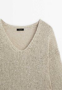 Beige, knitted sweater with a V-neck design. Features a loose weave texture and raw edges at the neckline and hem. Label visible inside.