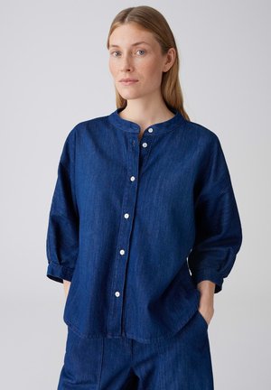 Frau mit heller Haut und blondem Haar trägt ein locker sitzendes dunkelblaues Jeanshemd mit Knöpfen und passende Hose, die Hände in den Taschen.