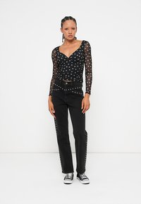 Frau mit geflochtenem Haar, die ein schwarzes, durchscheinendes Oberteil mit Punkten, schwarze jeans mit Nieten und schwarze Turnschuhe trägt und vor einem weißen Hintergrund steht.