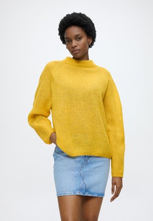 Žena s krátkymi kučeravými vlasmi v žltom pletenom svetri a svetlomodrej rifľovej mini sukni, stojaca s jednou rukou v vrecku.