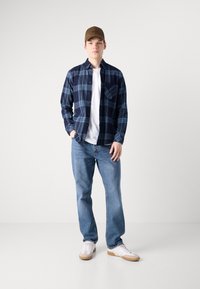 Camisa de botão xadrez azul-marinho e azul claro, com uma T-shirt branca por baixo, combinada com jeans azul claro e ténis brancos. Um boné castanho complementa o visual.