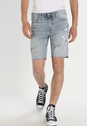 Homme marchant portant un short en denim bleu clair déchiré, un T-shirt blanc et des baskets montantes noires avec des lacets blancs.