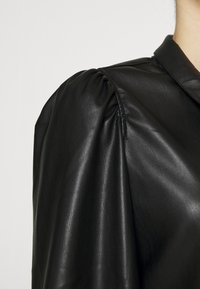 Blouse noire en cuir avec un col structuré et des détails bouffants aux épaules, arborant une texture lisse et un éclat subtil.