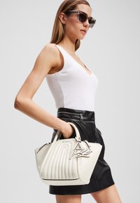 Sac à main blanc texturé avec un design à relief et un logo signature. Porte avec une jupe en cuir noire et un débardeur blanc. Lunettes de soleil présentes.