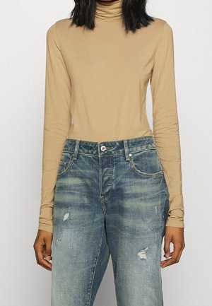 Femme portant un col roulé beige à manches longues rentré dans un jean bleu délavé taille haute avec une légère usure.