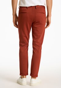 Rostfärgade byxor med skräddarsydd passform, tillverkade av mjukt material. Har två bakfickor och uppvikt i nederkant. Bärs med vita sneakers.