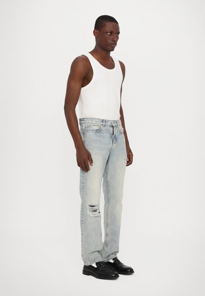 Falmet blå denimjeans med slidte detaljer, sat sammen med en hvid tanktop og sorte sko; model poserer i en neutral baggrund.