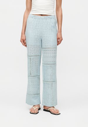 Kvinde iført lyseblå bukser med vide ben og geometriske blondecifre samt en hvid ærmeløs top, kombineret med beige thong-sandaler.