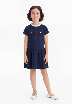 Mädchen mit kurzen dunklen Haaren steht und trägt ein marineblaues kurzärmeliges Kleid mit Knöpfen vorne und weiße Schuhe auf weißem Hintergrund.