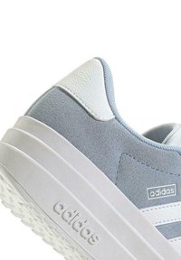 Sneaker in suede e pelle grigia con accenti bianchi, suola spessa e dettagli a tre strisce di lato. La marca è visibile sul tallone e sul lato.