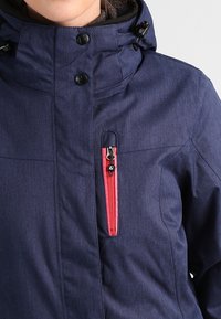 Marineblaue Outdoor-Jacke mit hohem Kragen, verstellbarem Kapuze und einer auffälligen roten Reißverschlusstasche auf der Brust, aus strukturiertem Material.