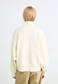 Felpa color crema con colletto alto, spalle calate e polsini a coste. Tessuto morbido con vestibilità rilassata, abbinata a pantaloni in velluto a coste beige.
