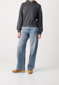 Sudadera de carbón con logo en el frente, combinada con vaqueros de denim azul claro de pierna ancha. Zapatillas amarillas-naranjas con acentos marrones completan el conjunto.