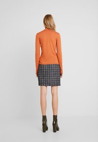 Pomarańczowy golf i spódnica w czarno-białe wzory, noszone z oliwkowymi botkami na obcasie; gładki dzianinowy materiał i teksturalne detale.