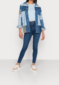 Chaqueta de mezclilla con diseño de patchwork en azul claro y oscuro; top ajustado azul claro; jeans oscuros ajustados; zapatillas beige y blancas.