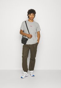 Tricou gri cu logo roșu, pantaloni cargo oliv și adidași albi cu accente albastre. Geantă de umăr neagră cu două închideri.