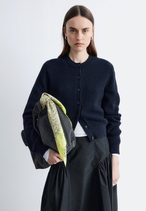 Vrouw met lang bruin haar, een marineblauw vest en een zwarte rok, die een zwarte tas vasthoudt met een gele bloemige sjaal, tegen een effen achtergrond.