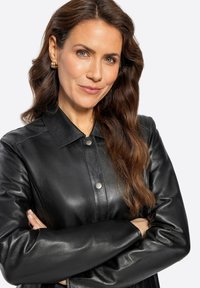 Chaqueta de camisa de cuero negro con collar, textura brillante, cierre frontal a presión y mangas largas, llevada por una mujer con cabello ondulado.