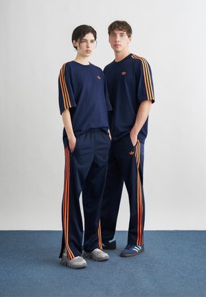 Δύο άτομα στέκονται δίπλα-δίπλα φορώντας πανομοιότυπες μπλε φόρμες με πορτοκαλί ρίγες και αθλητικά παπούτσια σε μπλε χαλί μπροστά σε λευκό τοίχο.