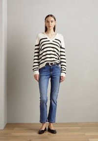 Stripet svart-hvit v-hals genser, kombinert med lyseblå jeans og svarte leiligheter, satt mot en nøytral bakgrunn.