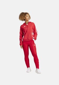 Tuta rossa con giacca con zip e cappuccio, strisce bianche sulle maniche e pantaloni aderenti. Realizzata in un tessuto sportivo e liscio.