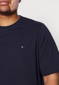 Mężczyzna w jednolitym ciemnoniebieskim t-shircie z małym prostokątnym logo w kolorach czerwonym, białym i niebieskim na piersi, na jednolitym tle.