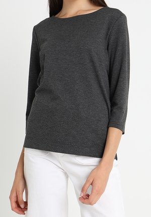 Femme portant un haut à manches longues gris foncé avec un col rond et un pantalon blanc, debout devant un fond blanc uni.