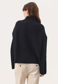 Pulover navy cu nervuri, cu guler înalt, croială lejeră și umeri căzuți, asortat cu pantaloni din fote deschis la culoare, având buzunare pe spate.