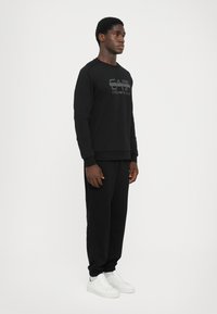 Svart hettegenser med teksturert logo, kombinert med matchende svarte joggebukser og hvite sneakers. Glatt stoff, avslappet passform, ribbede ermer.