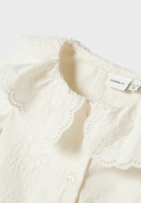 Blusa crema con colletto in pizzo scallop e ricami floreali. Presenta una chiusura a bottoni, tessuto morbido e dettagli intricati sull'orlo.