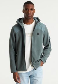 Hooded jack in donker teal met licht teal accenten, voorzien van een ritszak op de linkerarm en een gladde, gestructureerde stof.