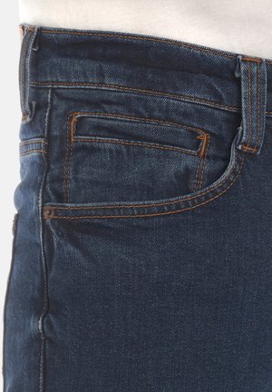 Granatowe dżinsy z jasnopomarańczowymi szwami, z tylną kieszenią i gładką fakturą. Góra kończy się w okolicach linii talii.