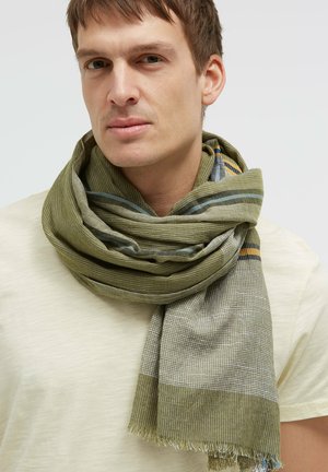 Young man wearing a large green and beige striped scarf wrapped around his neck over a light beige shirt.
