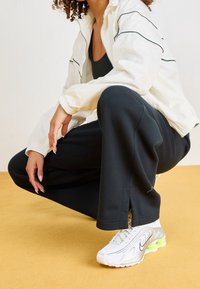 Bílá zipová bunda se zelenými pruhy, černé sportovní kalhoty a bílé tenisky s neonovými akcenty, na žlutém pozadí.