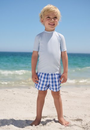 Fiatal gyermek mosolyog, világoskék napvédő felsőt és kék kockás rövidnadrágot visel, mezítláb áll a homokos parton, háttérben az óceán.