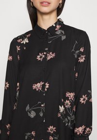 Nainen, jolla on päällään musta nappipusero, jossa on vaaleanpunaisia ja harmaita kukkakuoseja sekä kärjetön kaulus, yksinkertaisen vaalean taustan edessä.