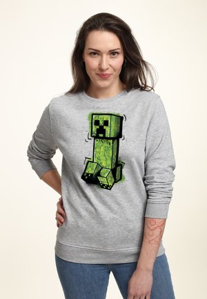 Szary sweatshirt z dużym zielonym, pikselowym grafiką Creepera na przodzie. Materiał wydaje się być miękki, z żebrowanymi mankietami i dołem.