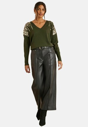 Olivgrüner V-Ausschnitt-Pullover mit metallischen Paillettenakzenten an den Schultern, kombiniert mit hochgeschnittenen schwarzen, weiten Hosen aus Kunstleder.