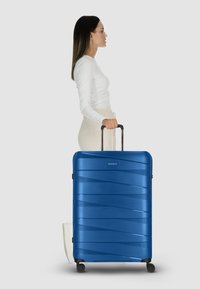 Maleta rígida azul con un diseño de olas texturizado, mango extensible y cuatro ruedas negras para una fácil movilidad.