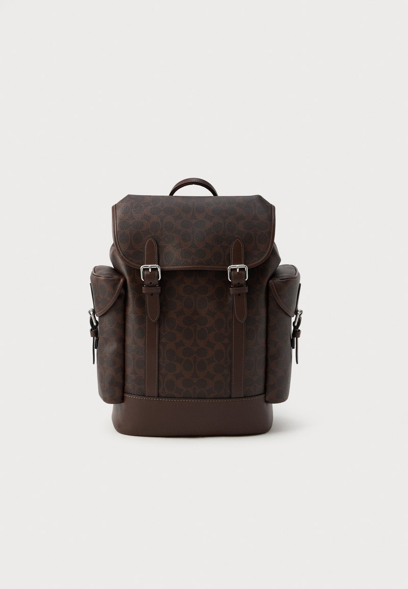 Zaino in finta pelle marrone con un esterno decorato, pattina anteriore con chiusura a doppia fibbia e tasche laterali con cinghie regolabili.